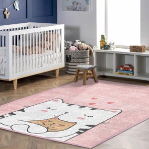 May include: A pink area rug with a cat design in a nursery setting. The rug features a large white cat hugging a smaller tan cat, with black and gray accents. Small red hearts are scattered around the cat design. A white crib and other nursery items are in the background.