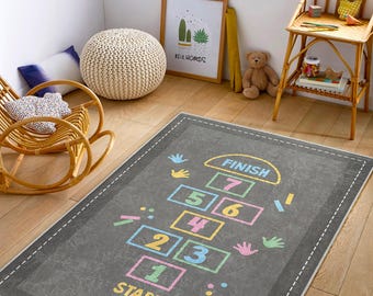 May include: A rectangular grey rug with a hopscotch design in pastel colours. The rug features the words "START" and "FINISH" and numbers 1 to 7. The rug is bordered with a white dashed line.