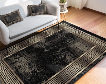 May include: A rectangular black area rug with a gold Greek key border. The rug has a distressed, textured appearance. The rug is placed on a hardwood floor, with a gray sofa and pillows in the background.