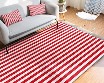 May include: A red and white striped rug is the focal point, with a grey sofa and matching striped cushions. A white wire chair and a small table with a plant are also in view. The rug's pattern is a series of bold stripes.