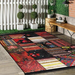May include: A rectangular patchwork rug with a black border. The rug features a variety of patterns and colors, including red, black, and floral designs. The rug is placed on a wooden floor.