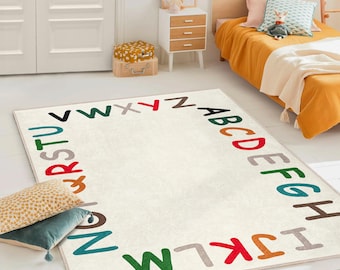 May include: A rectangular off-white rug with the alphabet in various colors around the border. The rug is in a child's bedroom, with a bed, nightstand, and decorative pillows visible.