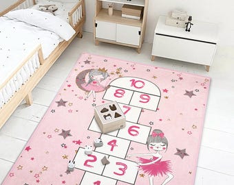 May include: A pink hopscotch rug with illustrations of ballerinas, stars, and the numbers 1-10. A wooden shape sorter and small animal figures are on the rug. The rug is in a child's bedroom with a bed and storage furniture.