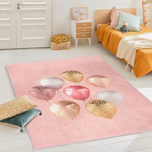 May include: A rectangular pink rug featuring a pattern of stylized balloons in gold, pink, and white. The balloons have a glittery or metallic appearance. The rug is designed for a child's room or nursery.