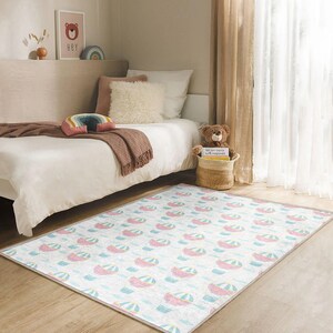 May include: A rectangular rug with a repeating pattern of pastel hot air balloons in pink, blue, and yellow on a white background. The rug is placed on a wooden floor in a bedroom setting. The rug's design adds a playful touch to the room's decor.