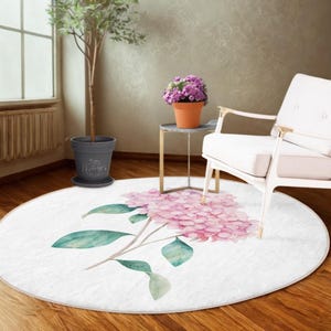 Puede incluir: Una alfombra redonda blanca con una ilustración de acuarela de una flor de hortensia rosa y hojas verdes. La alfombra está colocada sobre un suelo de madera, con una planta en maceta y una silla blanca en el fondo.