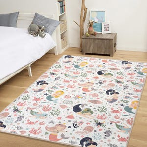 May include: A rectangular area rug with a white background featuring a colorful mermaid pattern. The design includes mermaids, sea creatures, coral, and starfish in shades of pink, blue, green, and yellow. The rug is placed on a wooden floor.