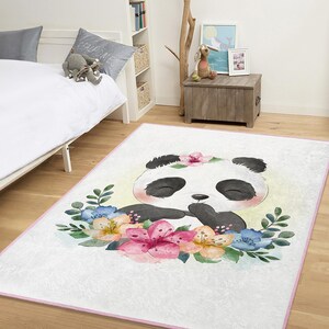 Op de afbeelding: Een rechthoekig wit tapijt met een panda-illustratie. De panda heeft zwart-witte vacht, gesloten ogen en een bloemenkrans. Kleurrijke bloemen en groene bladeren omringen de panda, met een roze rand.