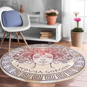 Peut inclure: Un tapis rond avec un fond beige et une illustration en bordeaux et noir du visage de Méduse avec des serpents pour cheveux. Le tapis a une bordure de clé grecque en noir et or.