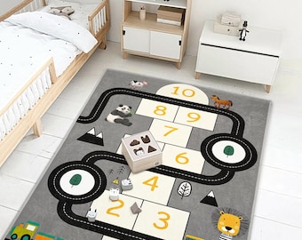 May include: A grey children's play rug with a hopscotch design, road graphics, and animal illustrations. The rug features a colourful train, a yellow school bus, and a lion. A wooden toy box with animal shapes sits on the rug.