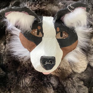 May include: A handmade dog mask with a black, white, and brown color scheme. The mask features white fur around the ears and face, with brown accents and mesh eye coverings. The dog's nose is black, and the tongue is pink.