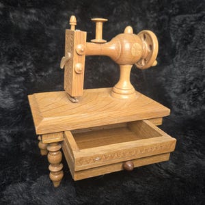 May include: A light brown wooden miniature sewing machine model featuring a flat table surface with a single pull-out drawer, a vertical needle bar, a top spool pin, and a side handwheel, all constructed from carved wood with visible grain patterns.