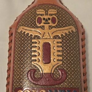 Könnte beinhalten: Eine braune Lederflasche mit einem goldenen und roten Design einer stilisierten Figur und dem Wort "COLOMBIA" in goldenen Buchstaben.