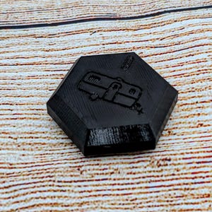 May include: A black hexagonal-shaped game piece with a raised image of a camper trailer.