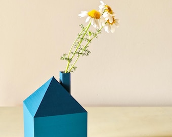May include: A teal blue house-shaped vase with a single stem of white daisies with yellow centers.