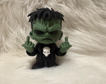 May include: A green figurine of Frankenstein's monster giving the middle finger with both hands. The figure has black hair, a black suit, and a white skull on the chest. The background is a fluffy white surface.
