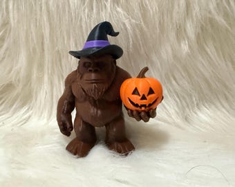 Puede incluir: Una figura marrón de Bigfoot con un sombrero de bruja negro con una banda morada, sosteniendo una calabaza naranja. La figura tiene una cara detallada y está posada sobre una superficie blanca y esponjosa. Decoración de Halloween.