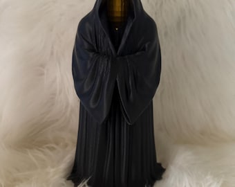 Könnte beinhalten: Eine schwarze Statue einer Kapuzenfigur mit einem drapierten Gewand. Die Figur hat ein goldenes Gesicht und steht auf einer weißen, flauschigen Oberfläche. Die Statue ist ein dunkles, dekoratives Objekt im gotischen Stil.
