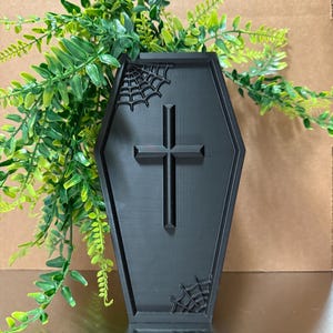 May include: Black coffin-shaped tray with a cross and spiderweb details. The tray is made of a solid black material and is decorated with a cross and spiderwebs. Green leafy plants are in the background.