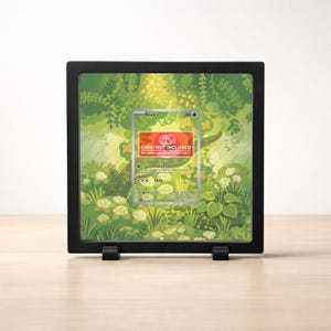 May include: A black display frame holding a Pokemon card with a green and white forest background. The card features a Rowlet character and the text "CARD NOT INCLUDED". The frame is on a light wood surface.