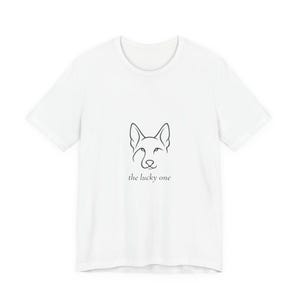 Puede incluir: Camiseta blanca con un dibujo lineal minimalista de la cara de un perro y el texto "the lucky one" en negro. El diseño está centrado, ofreciendo una estética limpia y moderna. Es una prenda de vestir informal.