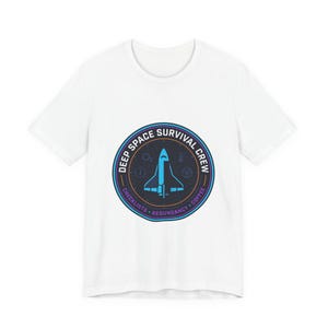 May include: White t-shirt featuring a circular graphic with a space shuttle design. The graphic includes the text "DEEP SPACE SURVIVAL CREW" and other words like "CHECKLISTS", "REDUNDANCY", and "COFFEE". The design is in shades of blue, purple, and white.