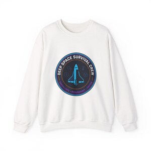 Peut inclure: Sweat-shirt blanc à col rond avec un motif circulaire. Le motif présente une navette spatiale bleue à l'intérieur d'un cercle bleu marine avec le texte "DEEP SPACE SURVIVAL CREW". Le texte supplémentaire comprend "CHECKLISTS", "REDUNDANCY" et "COFFEE".