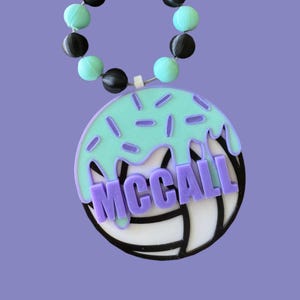 May include: A purple and white volleyball with green icing and sprinkles. The volleyball has the name "MCCALL" written on it. A black and turquoise beaded bracelet is attached to the top of the volleyball.