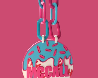 Pode incluir: Um colar com pingente com um design temático de beisebol. O pingente apresenta uma base de beisebol branca com costuras vermelhas, coberta com glacê azul e granulado rosa. A palavra "McCall" está em rosa. A corrente é rosa e azul.