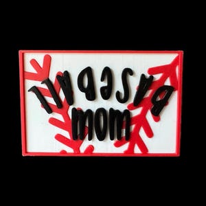 May include: A rectangular sign with a white background and a red border. The sign features the words "WASSA MOM" in black, with red snowflake-like designs. The overall design is simple and festive.