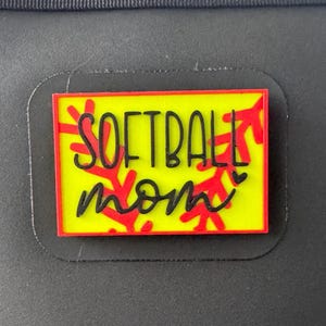 May include: A rectangular, yellow and red softball mom design. The design features the words "SOFTBALL mom" in black script, with red softball stitching accents. The design is set against a black background.