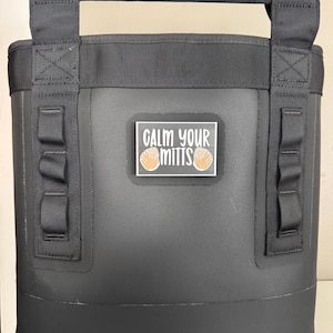 May include: Black insulated tote bag with a black handle and straps. The bag has a rectangular patch with the text "CALM YOUR MITTS" and two baseball mitts. The bag is designed to keep items cold.