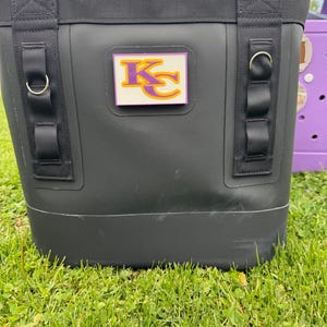 May include: Black insulated cooler bag with a purple and gold "KC" logo. The bag has black straps and metal D-rings. The cooler is sitting on green grass.