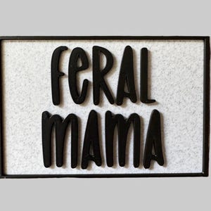 May include: A rectangular sign with the words "FERAL MAMA" in bold, black letters. The letters are raised against a white, textured background. The sign is framed with a thin, black border.