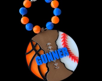 Könnte beinhalten: Ein brauner, weißer und orangefarbener Sport-Anhänger mit einer blauen Perlenkette. Der Anhänger hat ein Basketball-, Baseball- und Football-Design mit dem Namen "GUNNER" in Blau.