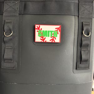 May include: A black insulated cooler bag with black straps and silver metal D-rings. A rectangular patch with the name "SMITH" in green letters is affixed to the front. Red baseball stitching and snowflake accents surround the name.