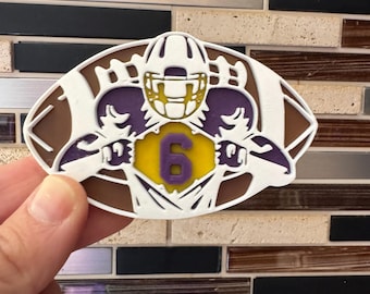 May include: A football-shaped emblem with a white outline, brown center, and purple and yellow accents. The design features a football helmet, two football players, and the number 6. The emblem is held in a hand.