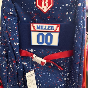 May include: A blue backpack with a white and red paint splatter pattern. It features a red B101 logo patch, a name patch reading MILLER, a number patch reading 00, and a red horizontal strap with a white plastic buckle across the front.