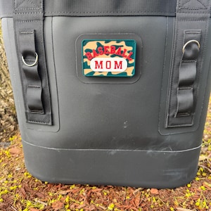 May include: Black insulated tote bag with a "BASEBALL MOM" patch. The patch has a camouflage background with red and white lettering. The bag has black straps and metal D-rings.