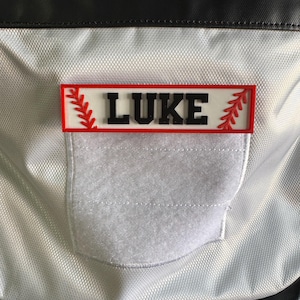 May include: A baseball-themed patch with the name "LUKE" in black block letters. The patch has a red border and red baseball stitching accents. It is attached to a silver and black bag with a white velcro patch.