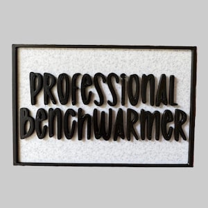 May include: A rectangular sign with a black frame and a white textured background. The words "PROFESSIONAL Benchwarmer" are in bold, black, 3D lettering. The sign is a humorous decorative item.