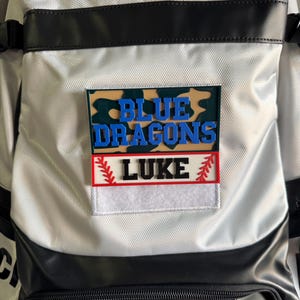 May include: A white and black baseball bag with a patch that says "BLUE DRAGONS" in blue letters over a camouflage background. Below, a red and white patch reads "LUKE".