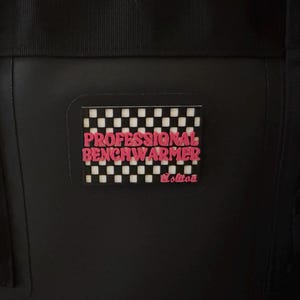 May include: Black bag with a rectangular patch featuring the words "PROFESSIONAL BENCHWARMER" in pink, set against a black and white checkered background. The bag has black straps and a textured surface.
