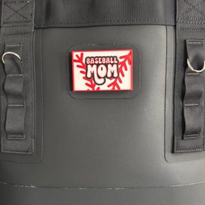 May include: A black cooler bag with a rectangular patch that reads "BASEBALL MOM" in red and white. The bag has black straps and silver metal rings.