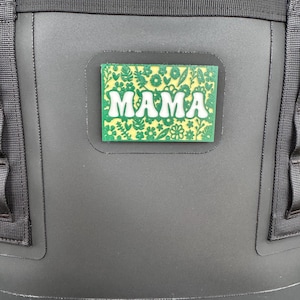May include: A rectangular patch with the word "MAMA" in white, stylized letters. The patch has a green background with a floral pattern. The patch is affixed to a black bag with black straps.