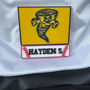 May include: A yellow and navy blue patch featuring a cartoon tornado with a smiling face. Below the tornado is a rectangular patch with the name "HAYDEN S" in black letters, with red and white baseball-themed accents.