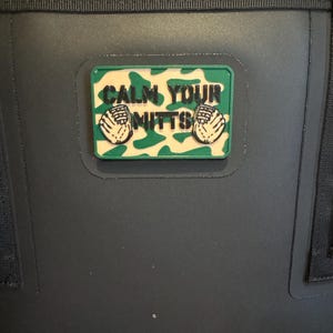 May include: A rectangular patch with the text "CALM YOUR MITTS" in black lettering. The patch has a green and tan camouflage background and two white baseball mitts. The patch is attached to a dark gray surface.
