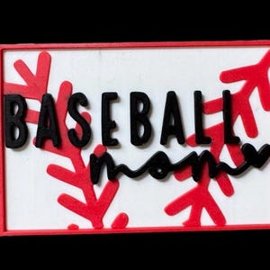 May include: A rectangular sign with a white background and a red border. The words "BASEBALL mom" are in black, with red decorative accents. The sign is likely a decorative item.