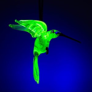 May include: A neon green glass hummingbird ornament with outstretched wings, suspended against a dark blue background. The bird has a long, black beak and a detailed tail. The vibrant green color glows, creating a striking contrast.