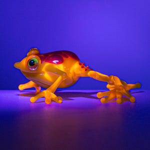 Puede incluir: Una pequeña figura de rana amarilla y naranja con manchas rojas y ojos verdes. La rana está posando con una pata extendida, sobre un fondo azul degradado. La figura probablemente está hecha de vidrio o un material similar.